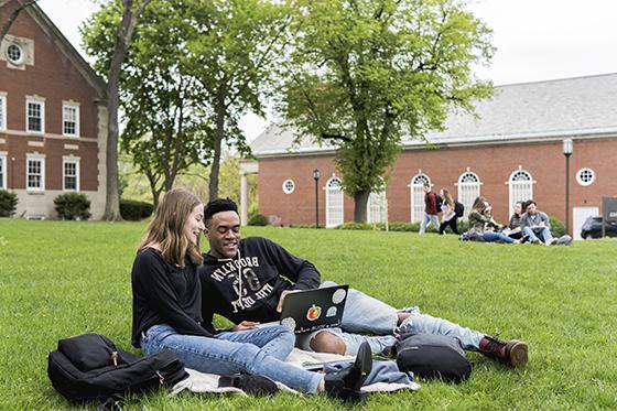 两名波胆网站的学生坐在绿草地上的学术广场上，一起用笔记本电脑工作. 