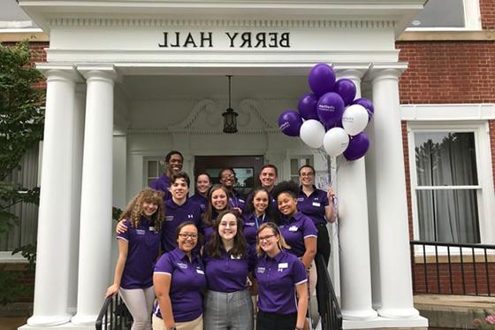 Photo of a group of Chatham University 工作人员 st和ing in front of Berry Hall wearing purple shirts 和 holding purple balloons. 