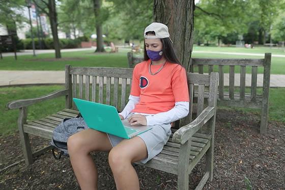 波胆网站学生戴面具的照片, 外, 坐在长凳上摆弄她的电脑.